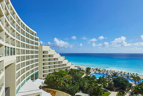 A modern beachfront resort hotel in Mexico with balconies overlooks a turquoise ocean, sandy beach, palm trees, and a large outdoor pool—perfect for meetings or conference events under a bright blue sky with scattered clouds.