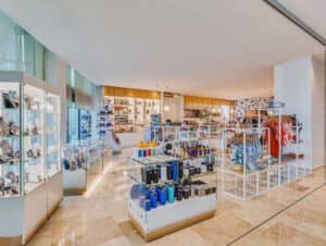 Una tienda de regalos moderna y luminosa en los complejos turísticos de México, con vitrinas de cristal que exhiben joyas y accesorios, además de estantes con coloridas botellas de agua, trajes de baño y recuerdos; perfecta para eventos o reuniones sobre un suelo de baldosas pulidas.