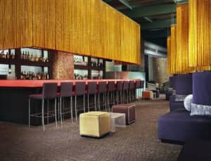 El interior de un bar moderno en los complejos turísticos de México cuenta con una larga barra con sillas moradas, estantes con botellas de licor detrás, cortinas de cadenas doradas que cuelgan del techo y otomanas cúbicas sobre una alfombra estampada; ideal para reuniones o eventos de conferencias.