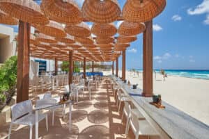 Un restaurante al aire libre junto a la playa en los complejos turísticos de México, con sillas blancas, mesas de madera y lámparas de techo con forma de cesta tejida, perfecto para eventos de conferencias o reuniones relajadas en la arena con impresionantes vistas al océano y un cielo azul de fondo.