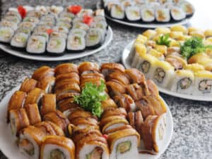 Cuatro platos con surtidos de rollos de sushi se exhiben sobre una encimera de granito, ideal para resorts en México o eventos de conferencias. El plato en primer plano presenta rollos cubiertos con aguacate en rodajas y salsa, adornados con perejil, mientras que los demás ofrecen variedades coloridas.