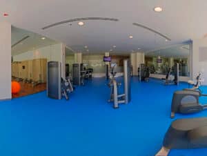 Un gimnasio moderno en uno de los resorts más exclusivos de México, con piso azul, diversas máquinas de ejercicio y grandes espejos. A la izquierda se encuentran los casilleros y una pelota de ejercicio naranja, lo que hace de este espacio bien iluminado un lugar ideal para los huéspedes que asisten a conferencias o reuniones.