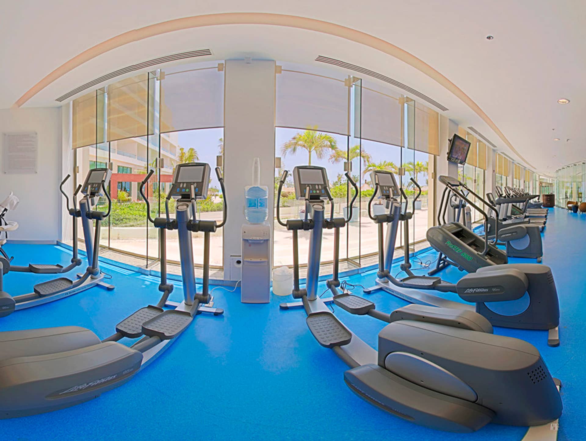 A modern gym in Mexico resorts with blue flooring, elliptical machines, treadmills, and a water dispenser—all facing large windows with views of palm trees and a sunny outdoor area. Ideal for guests attending meetings or conference events.