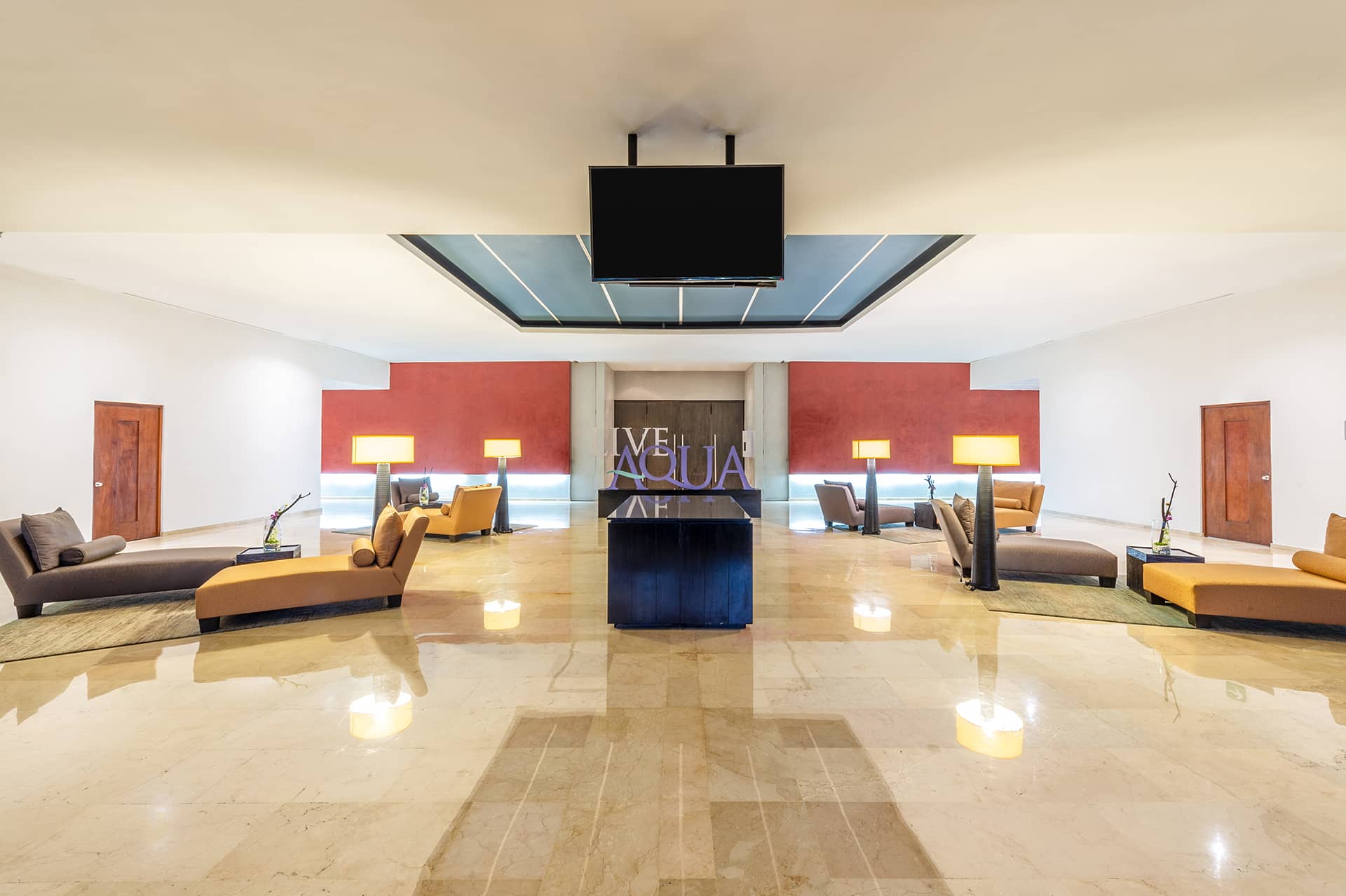 Spacious, modern hotel lobby with polished marble floors, tan and brown lounge chairs, floor lamps, and decorative plants. Ideal for conference events or meetings, this Mexico resort features LIVE AQUA signage and a central black table beneath a ceiling TV.