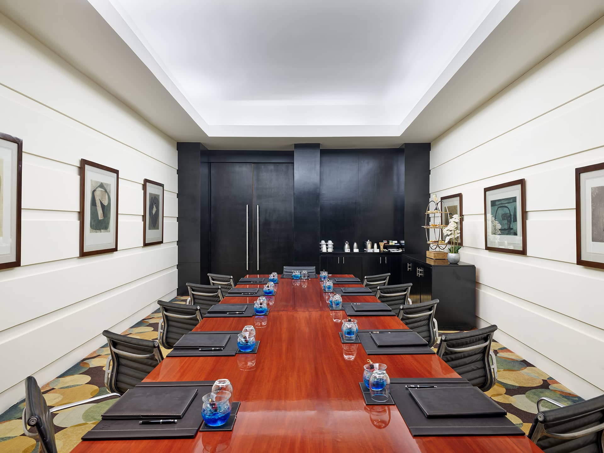 A modern, well-lit conference room in one of Mexico's finest resorts, featuring a long wooden table, black chairs, notepads, pens, and water glasses—ideal for meetings or conference events. Framed artwork and a refreshment station complete the space.