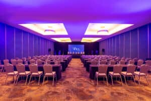 Una amplia y moderna sala de conferencias en un resort mexicano, con filas de sillas y mesas frente a un escenario con pantalla de proyección, iluminada con luces de techo moradas y amarillas. El espacio es ideal para reuniones de hotel, presentaciones o eventos de conferencias.