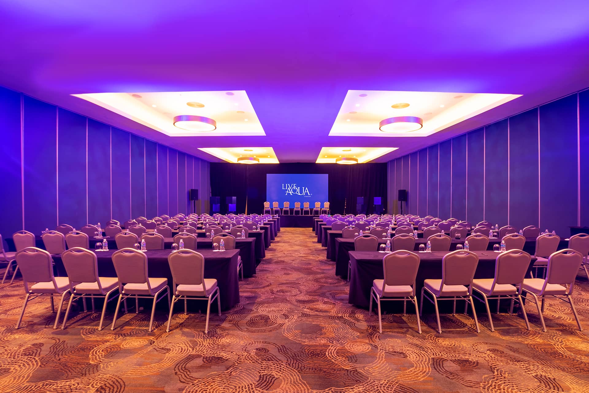A large, modern conference room in a Mexico resort with rows of chairs and tables facing a stage with a projection screen, lit by purple and yellow ceiling lights. The space is ideal for hotel meetings, presentations, or conference events.