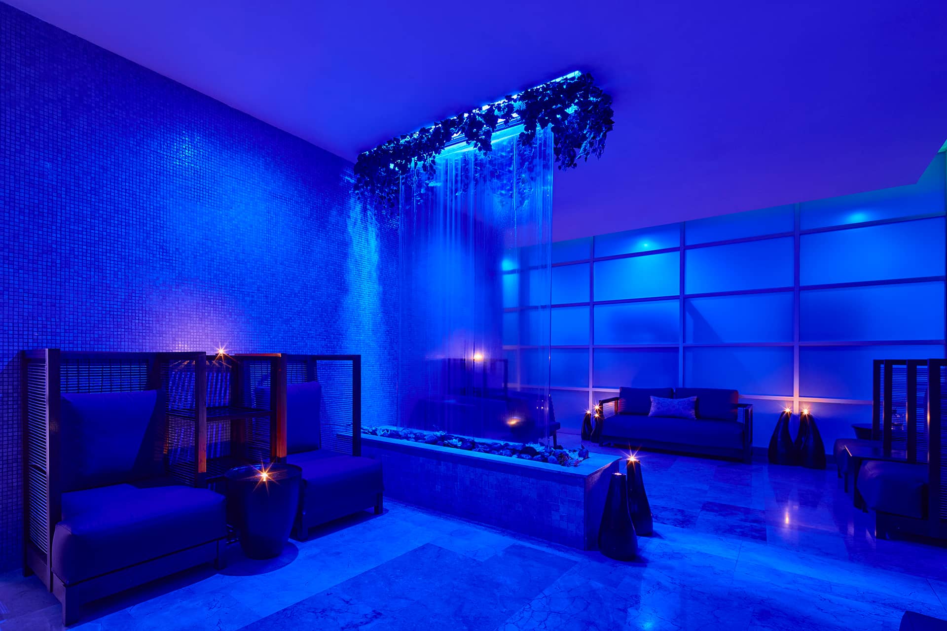 A modern lounge with blue mood lighting, comfortable seating, small tables with candles, and a central water feature with cascading water and hanging greenery—perfect for relaxing between meetings at Mexico resorts or conference events.