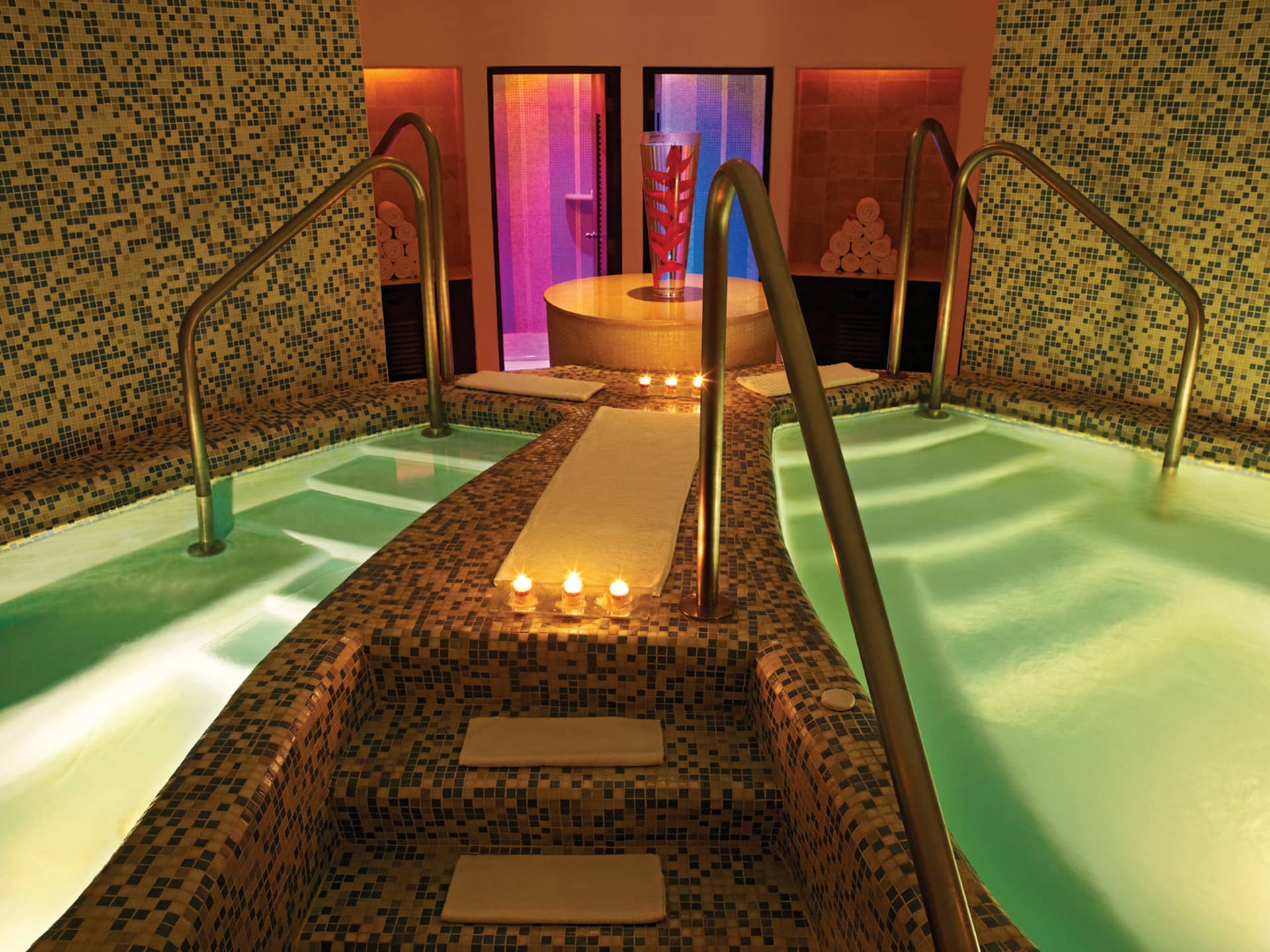 A luxurious spa room in one of Mexico's finest resorts, with two mosaic-tiled hot tubs, soft lighting, and candles, leading to a central soaking tub in front of a glass door glowing with pink and orange lights—ideal for conference events or relaxing after meetings.
