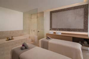 Una moderna sala de spa en uno de los mejores resorts de México, que cuenta con dos camillas de masaje con sábanas de color beige, toallas enrolladas con flores, una ducha con mampara de cristal, lavabos dobles e iluminación ambiental suave para crear un ambiente relajante entre los eventos de la conferencia.