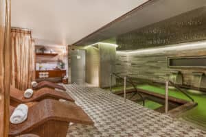 Una moderna sala de spa en uno de los mejores resorts de México cuenta con tumbonas de mimbre, toallas enrolladas, suelo de baldosas, iluminación tenue y una piscina climatizada con cascadas junto a una pared de piedra, perfecta para relajarse después de reuniones o eventos de conferencias.