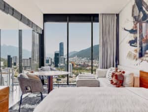 Habitación de hotel moderna con cama, sofá, mesa y sillas, con grandes ventanales que ofrecen vistas al horizonte de la ciudad y las montañas. Ideal para reuniones o conferencias, este hotel en México cuenta con luz natural y llamativas obras de arte abstracto en las paredes.