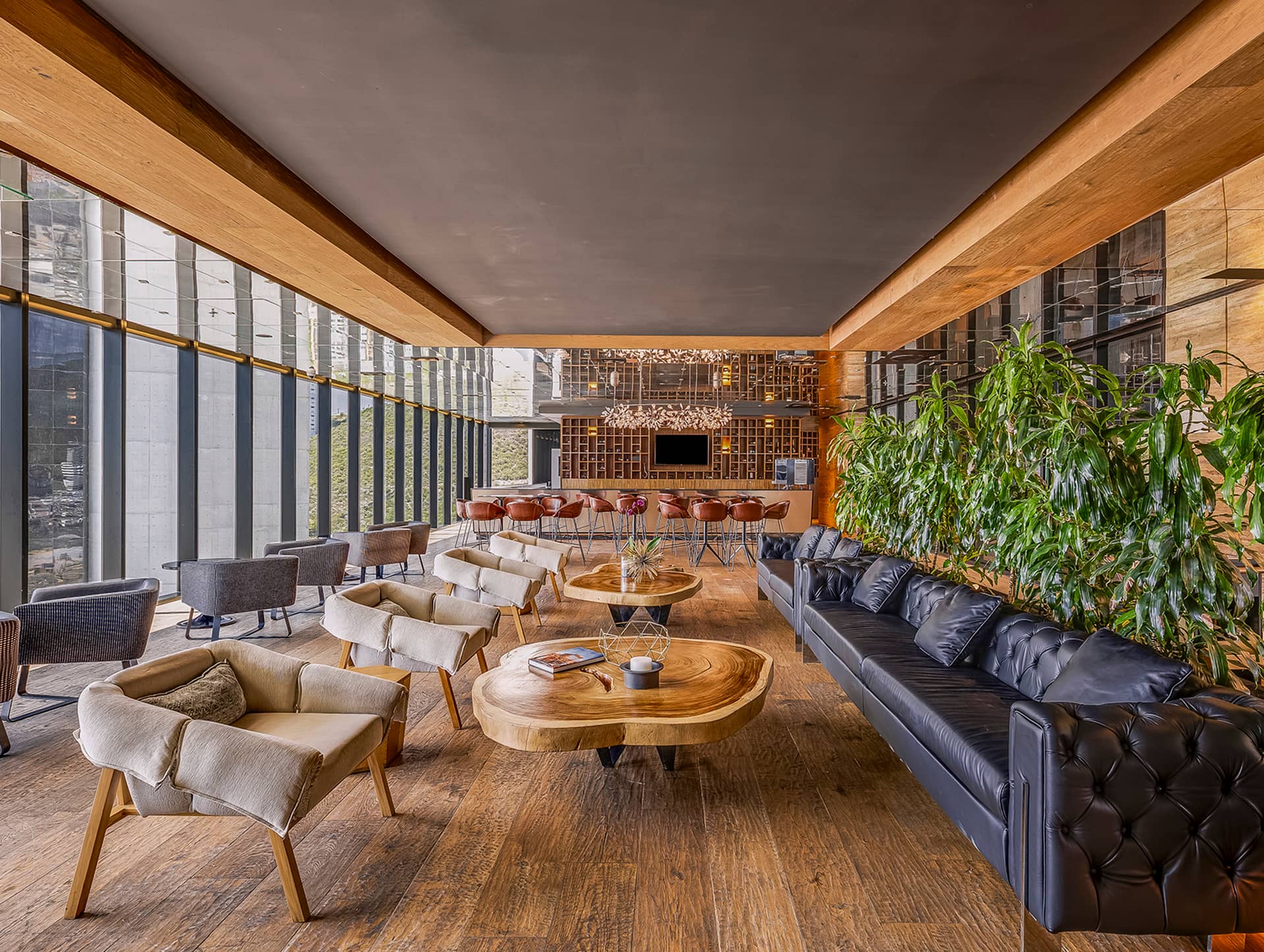 A modern lounge in Mexico resorts with wooden floors, a black leather sofa, cream chairs, round wooden tables, and tall green plants. Large floor-to-ceiling windows and a bar area make it ideal for meetings or conference events.