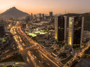 Un paisaje urbano al atardecer con altos edificios modernos, calles iluminadas y una montaña al fondo. La escena es vibrante, con muchos coches y luces de la ciudad que empiezan a brillar al caer la noche; ideal para conferencias o reuniones.