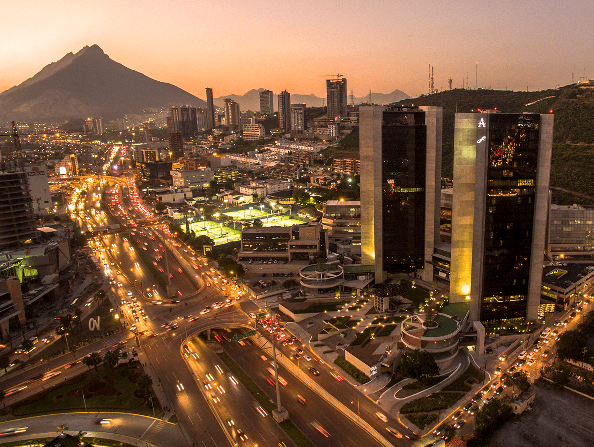A cityscape at sunset with tall modern buildings, busy illuminated roads, and a mountain in the background. The scene is vibrant, with many cars and city lights starting to twinkle as evening falls—perfect for conference events or meetings.