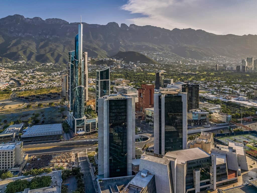 Aerial view of a modern city skyline with tall glass skyscrapers, surrounded by green areas and mountains—an ideal backdrop for conference events and meetings at top Mexico hotels and resorts under a partly cloudy sky.