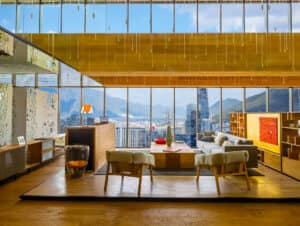 Una sala de estar moderna y elegante con grandes ventanales que ofrecen impresionantes vistas a la ciudad y a las montañas, ideal para relajarse después de eventos de conferencias en hoteles o resorts de México. Muebles en tonos neutros, detalles en madera, obras de arte en rojo y lámparas colgantes completan este acogedor espacio.