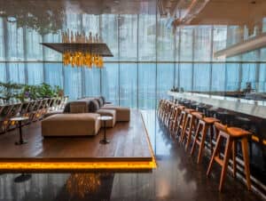 Moderna zona de estar y bar con elegantes sillas de madera en una larga barra, sofá central sobre una plataforma elevada, iluminación cálida, vegetación y grandes ventanales: ideal para eventos o reuniones en complejos turísticos y hoteles de México.