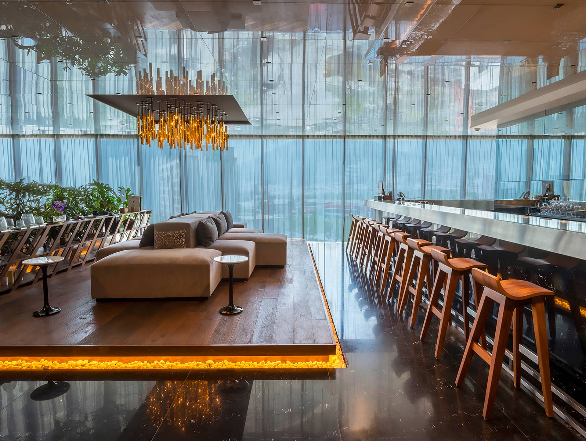 Modern lounge and bar area with stylish wooden chairs at a long counter, central sofa on a raised platform, warm lighting, greenery, and large windows—ideal for conference events or meetings at resorts and hotels in Mexico.