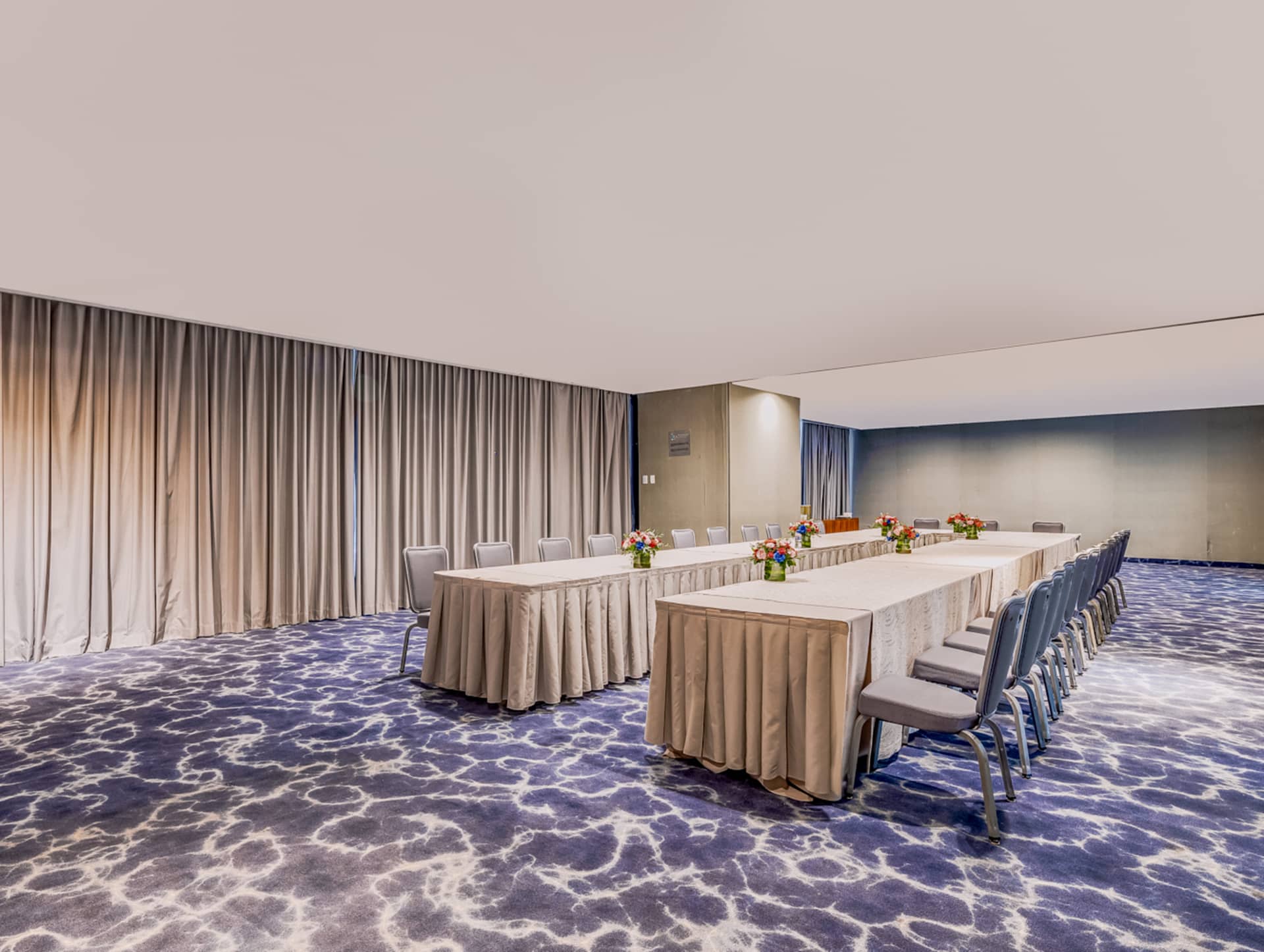 A modern conference room ideal for meetings and conference events, with U-shaped tables covered in beige tablecloths, gray chairs, and floral centerpieces. The space features gray curtains and a blue-and-white abstract patterned carpet.