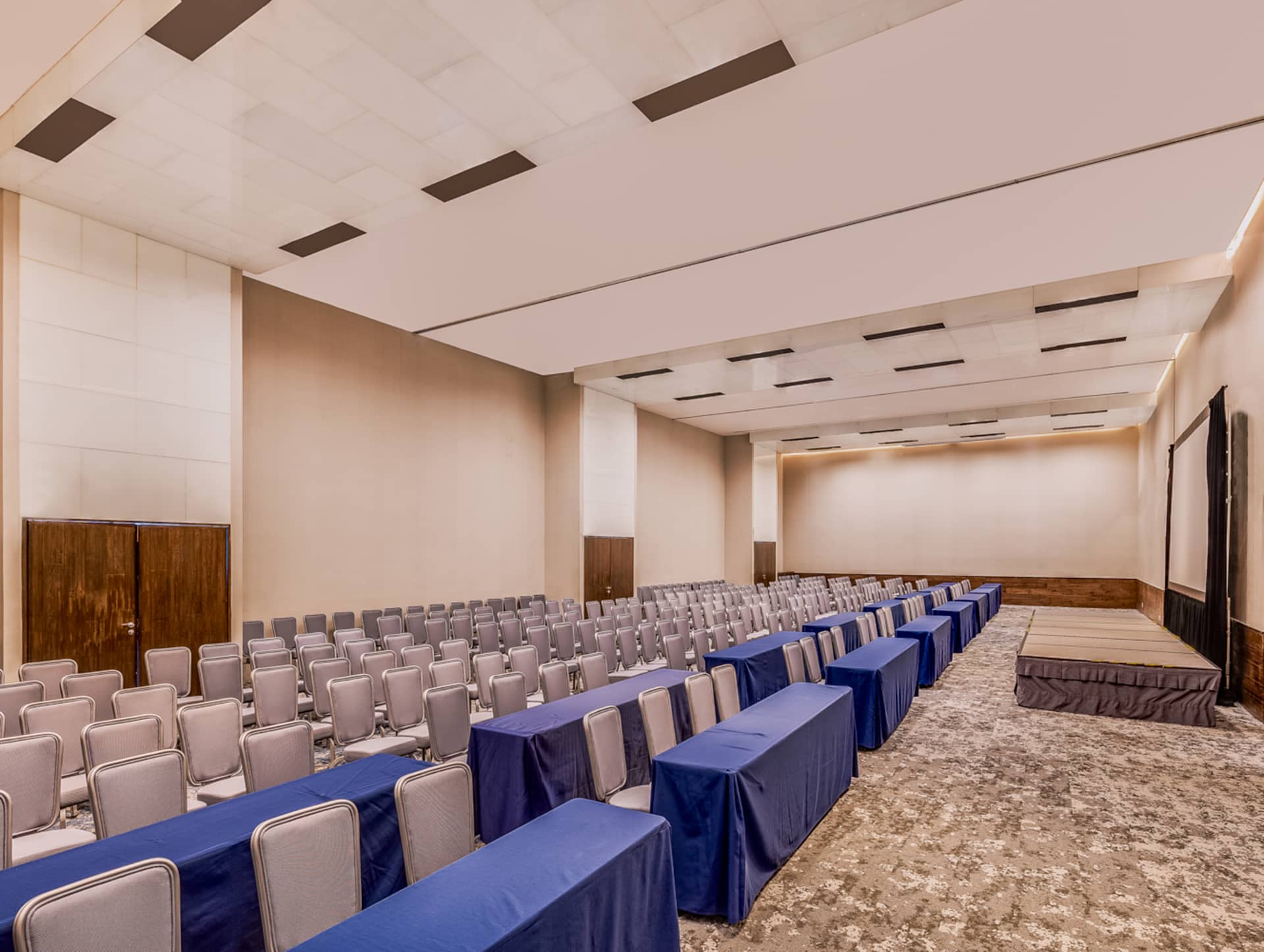 A large, empty conference room at a Mexico resort with rows of gray chairs facing a small stage and tables covered with blue tablecloths—ideal for meetings and conference events. The room features neutral walls, a patterned carpet, and ceiling lights.