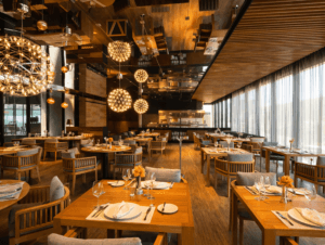 Un restaurante amplio y moderno en uno de los mejores complejos turísticos de México, con mesas y sillas de madera, grandes ventanales con cortinas transparentes, elegantes lámparas colgantes esféricas y mesas cuidadosamente dispuestas, ideal para reuniones o eventos de conferencias en un ambiente cálido y acogedor.