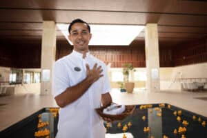Un miembro del personal del hotel, sonriente y con uniforme blanco, se encuentra en el interior, junto a un estanque reflectante con patitos de goma amarillos flotando, sosteniendo una cesta con toallas; una imagen que refleja la cálida hospitalidad de los hoteles mexicanos, ideales para reuniones y eventos de conferencias.
