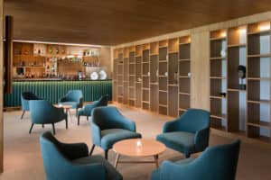 Salón moderno ideal para reuniones, con sillones color turquesa y mesas redondas de madera, una barra iluminada con estantes para botellas y cristalería, además de una gran pared con estantes abiertos de madera. La iluminación cálida crea un ambiente acogedor, perfecto para eventos y conferencias.