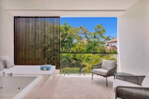 Un balcón moderno en uno de los resorts más exclusivos de México cuenta con una bañera integrada, dos sillas acolchadas y una mampara de listones de madera, que ofrece vistas serenas de frondosos árboles, perfectas para relajarse después de eventos o reuniones de conferencias.