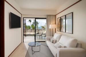 Un moderno y luminoso salón en un complejo turístico de México cuenta con un sofá blanco, una mesa de centro, un televisor montado en la pared y grandes puertas de cristal que dan a un patio con muebles de exterior, palmeras y vistas al mar, perfecto para relajarse después de los eventos de la conferencia.