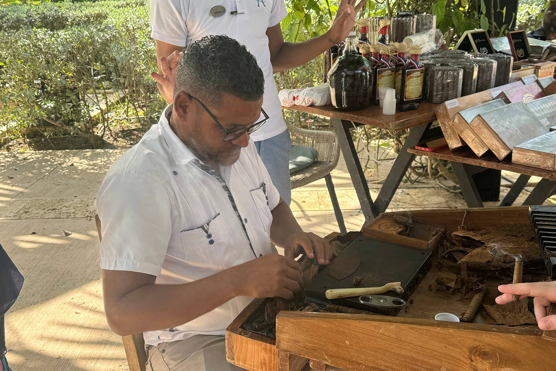 A man sits at a wooden table, skillfully rolling cigars by hand. Tobacco leaves and tools are spread out in front of him. Another person stands nearby, partially out of frame, at an outdoor market or Mexico resort workshop.