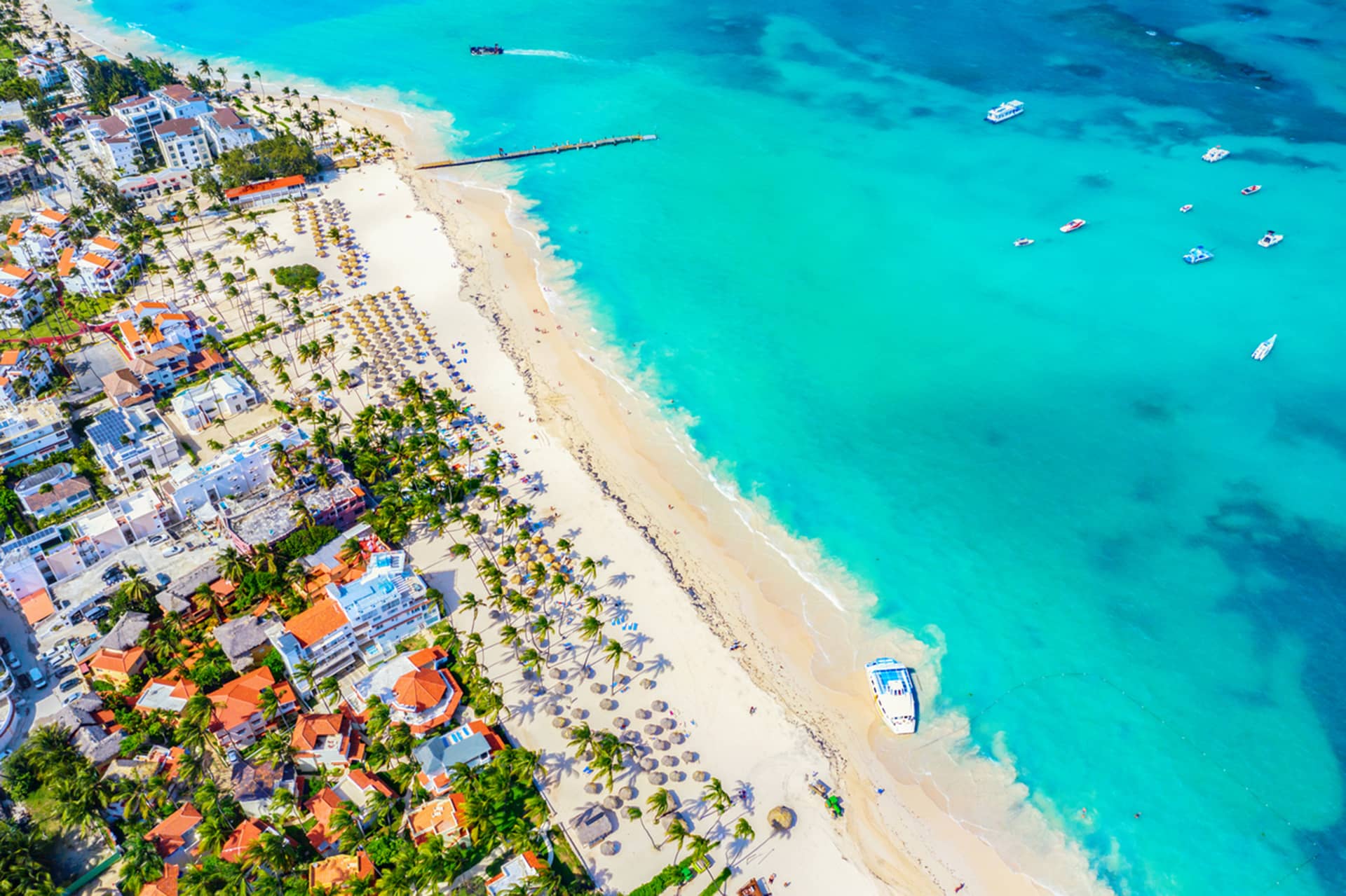 Aerial view of a tropical beach in Mexico with turquoise water, white sand, palm trees, and resort buildings. Boats are visible in the ocean, and rows of umbrellas line the beach—perfect for conference events and meetings at luxury hotels.