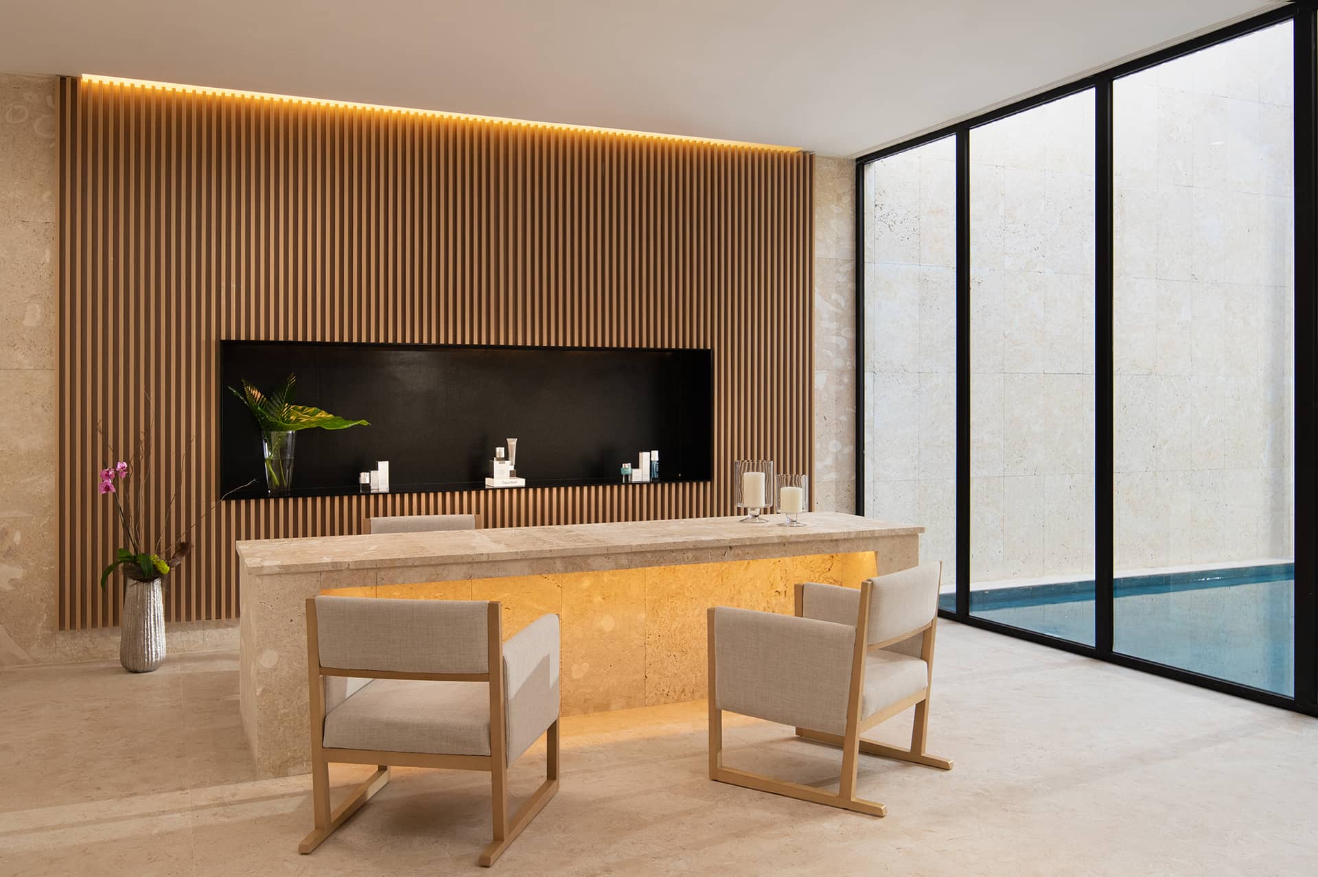 A modern spa room in one of Mexico's top resorts, featuring a marble counter, two beige chairs, a wooden slat accent wall, and floor-to-ceiling windows overlooking the pool. A purple orchid adds elegance to this well-lit, minimalist space.