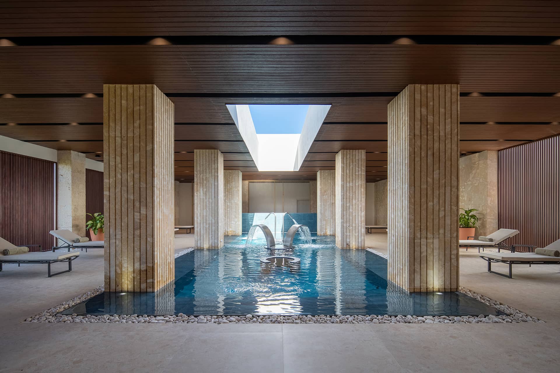 Luxurious indoor pool with four stone pillars, wooden ceiling, skylight above, poolside loungers with towels, and elegant lighting—perfect for relaxing after conference events or meetings at upscale Mexico hotels and resorts.