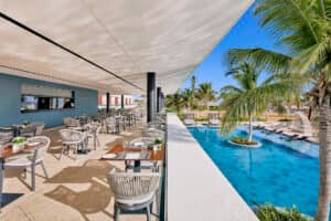 Restaurante al aire libre con mesas dispuestas junto a una piscina de aguas cristalinas, rodeado de palmeras y tumbonas bajo un cielo brillante y soleado: un entorno ideal para eventos de conferencias o reuniones informales en los mejores complejos turísticos de México.