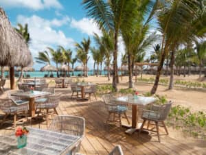 Restaurante al aire libre junto a la playa, con mesas y sillas de mimbre vacías sobre una terraza de madera, ideal para reuniones informales o eventos de conferencias, rodeado de palmeras y con vistas a las playas de arena en uno de los mejores resorts de México. Pequeños jarrones con flores decoran las mesas.
