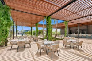 Terraza exterior con modernas sillas de mimbre y mesas redondas, ideal para eventos o reuniones. Una pérgola de madera con enredaderas proporciona sombra, y bajo el cielo azul de México se divisa una zona de bar.