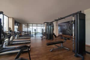 Gimnasio moderno con suelo de madera en este resort mexicano, equipado con cintas de correr, bicicletas estáticas, bancos de pesas y un mural con temática deportiva, ideal para huéspedes que asisten a conferencias o reuniones. Grandes ventanales inundan el espacio de luz natural.