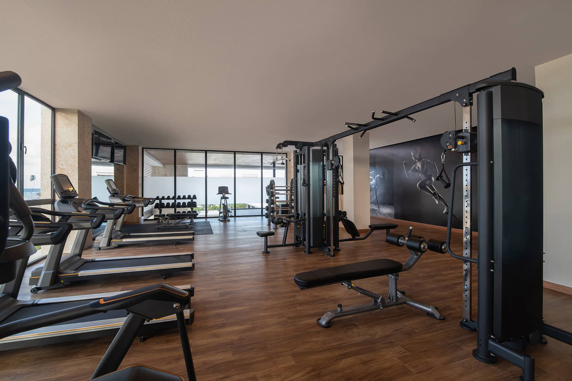 Modern gym with wood flooring at this Mexico resort, featuring treadmills, stationary bikes, weight benches, and a fitness-themed mural—perfect for guests attending conferences or meetings. Large windows fill the space with natural light.