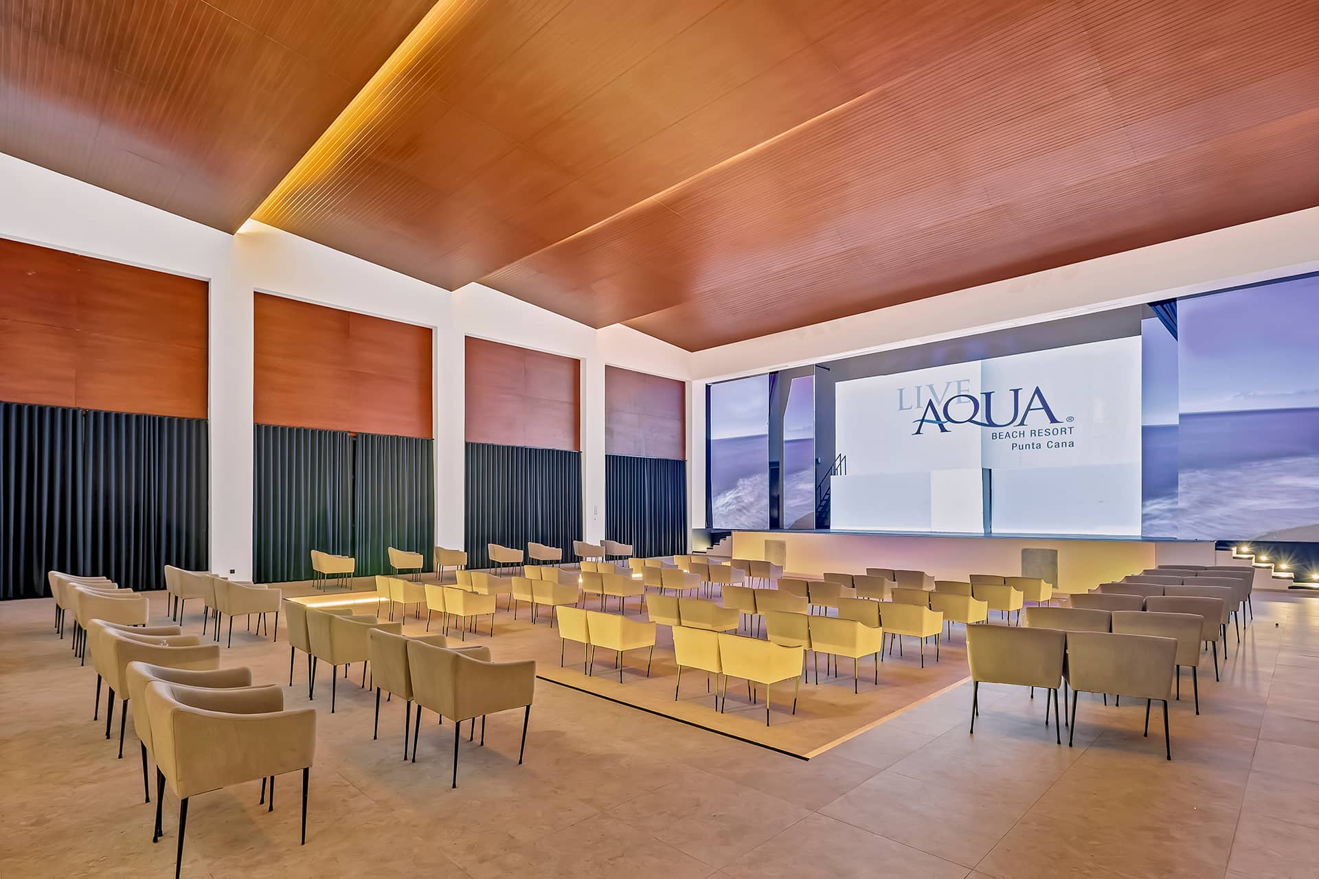 Modern conference room with rows of tan chairs facing a stage and large screen displaying Live Aqua Beach Resort Punta Cana, ideal for conference events or meetings, featuring a wood panel ceiling and large windows like top resorts and hotels in Mexico.