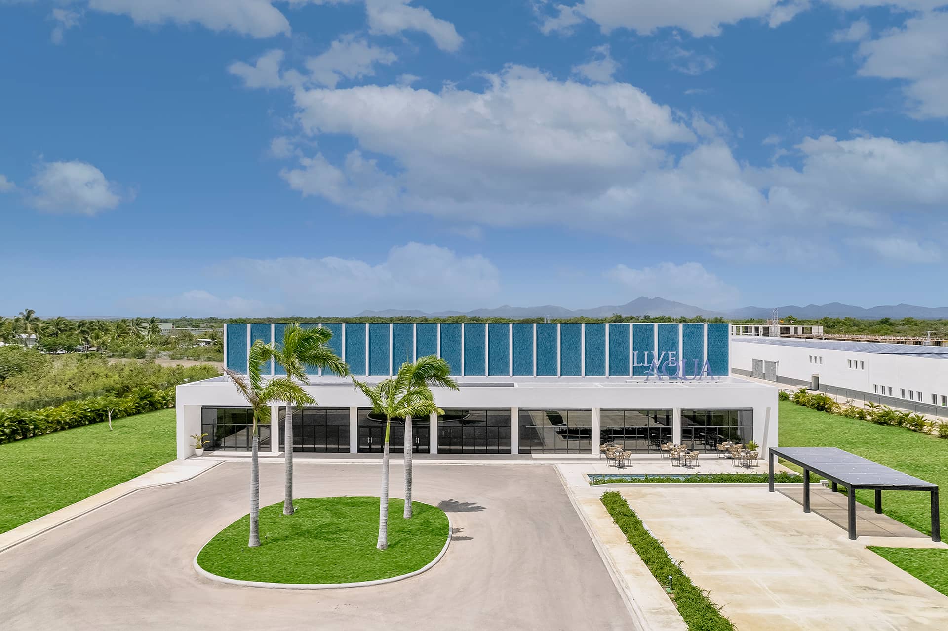 Modern, glass-fronted resort hotel with blue accents, surrounded by green lawns and palm trees under a partly cloudy sky. A covered parking area is visible on the right, with outdoor seating near the entrance—ideal for meetings or conference events.