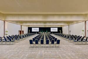 Una amplia y moderna sala de conferencias, ideal para reuniones y eventos, con filas de sillas azules frente a un escenario con la imagen de AQUA. La paleta de colores neutros y los arreglos florales le aportan elegancia, perfecta para hoteles y resorts en México.