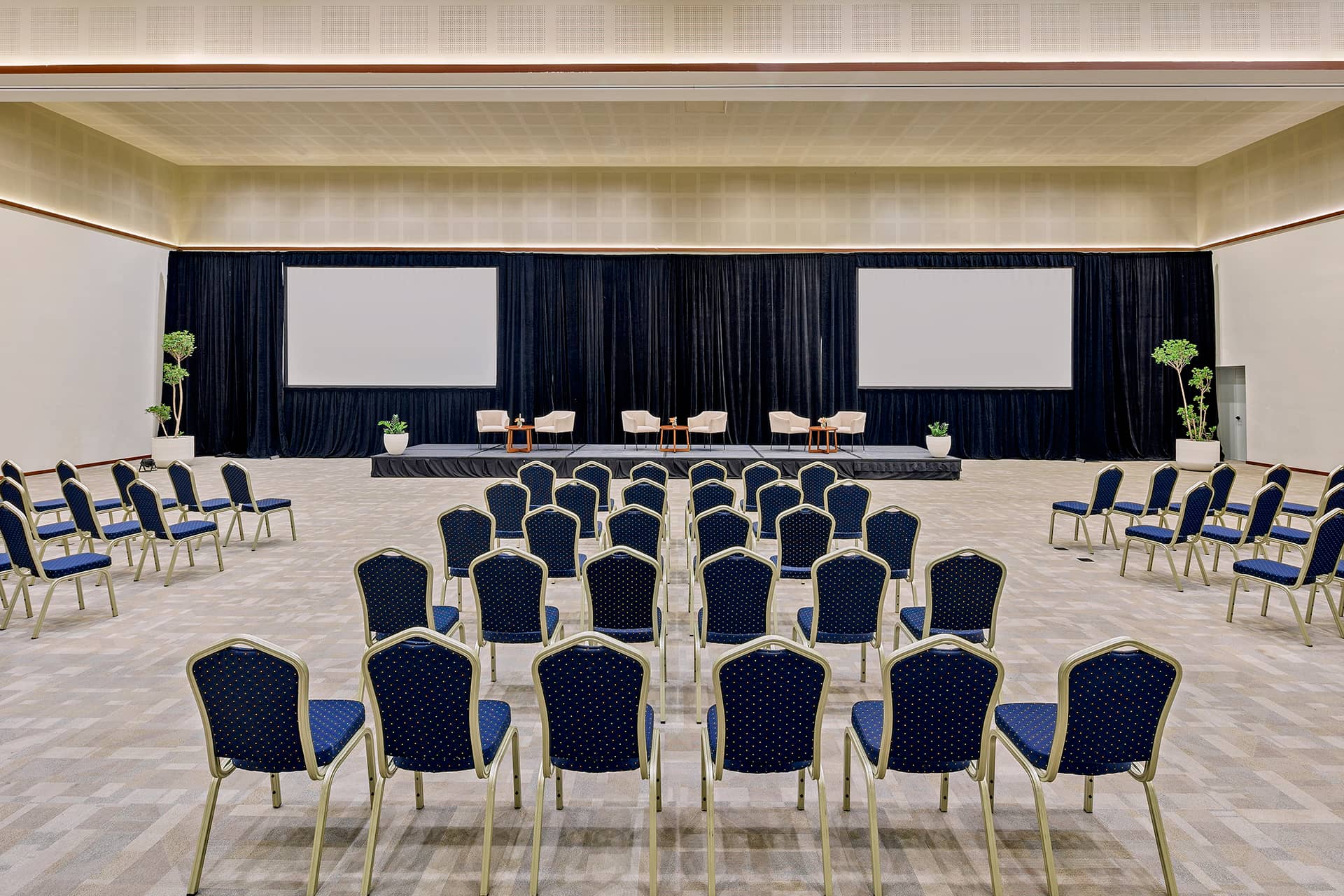 Rows of blue chairs face a stage with four armchairs, small tables, plants, and two large projection screens in a spacious, modern conference hall—ideal for meetings or conference events at top Mexico hotels and resorts.