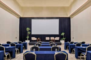 Una sala de conferencias en un complejo turístico de México con filas de mesas y sillas cubiertas con manteles azules, frente a un escenario con tres sillones, mesas pequeñas, plantas en macetas y una gran pantalla de proyección; ideal para reuniones y eventos de conferencias.