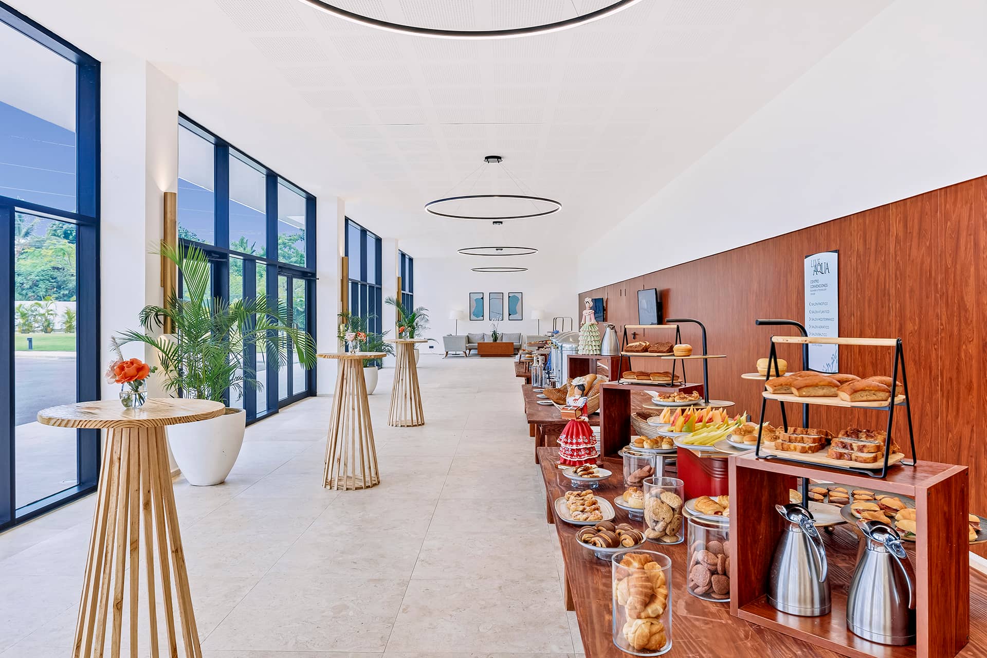 A bright, modern buffet area at this Mexico resort features tables with various pastries, fruits, and drinks. Large windows let in natural light, while tall potted plants add greenery—perfect for conference events or hotel meetings.