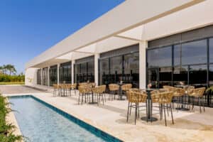 Los modernos patios al aire libre de los complejos turísticos mexicanos cuentan con sillas de mimbre y mesas redondas junto a un estanque reflectante, perfecto para eventos o reuniones, ubicado al lado de un edificio blanco con grandes ventanales bajo un cielo azul despejado.