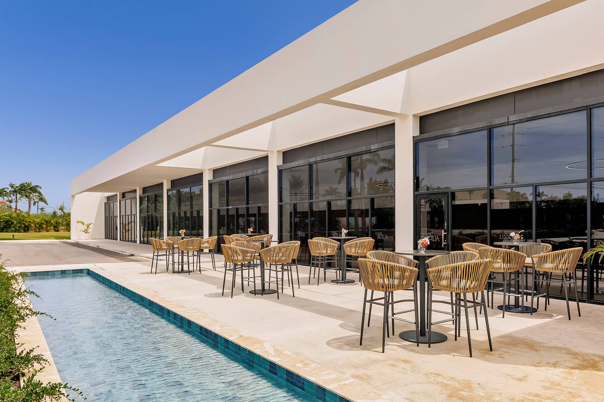 Modern outdoor patio at Mexico resorts features wicker chairs and round tables by a reflecting pool, perfect for conference events or meetings, set next to a white building with large glass windows under a clear blue sky.