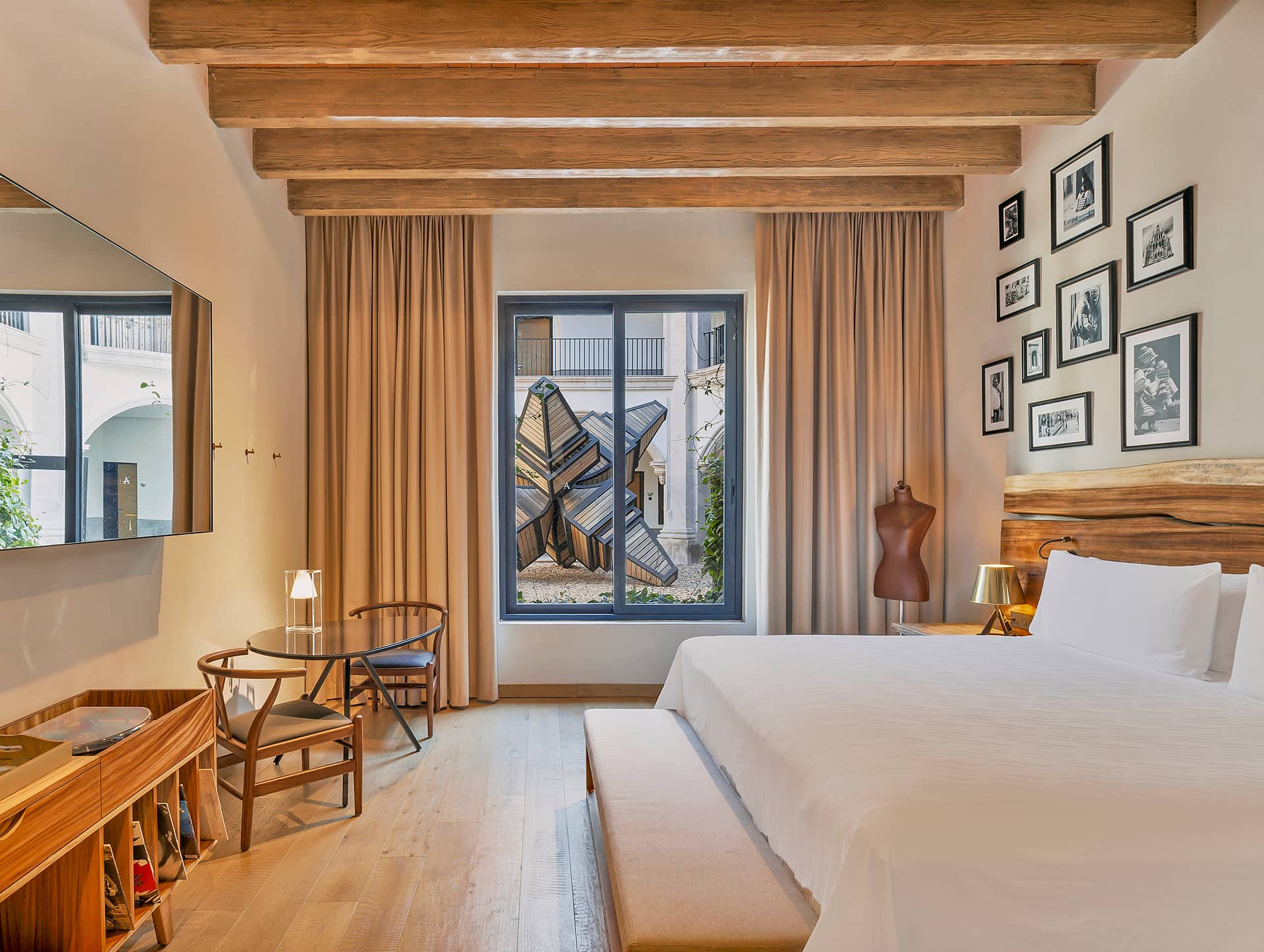 A modern hotel room with a large bed, wooden beams, a sitting area for meetings, wall art, and a window with beige curtains overlooking a courtyard featuring a unique metal sculpture. Perfect for guests attending conference events.