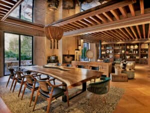 Un espacioso comedor, ideal para reuniones, cuenta con una gran mesa de madera con borde natural y sillas modernas, una lámpara de araña tejida, vigas de madera a la vista, estanterías empotradas con elementos decorativos y grandes ventanales que dejan entrar la luz natural.