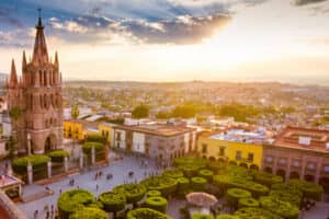 Vista aérea de una plaza histórica con una gran iglesia neogótica, árboles verdes bien cuidados, coloridos edificios coloniales y gente paseando al atardecer cerca de los mejores hoteles y centros de convenciones de México.
